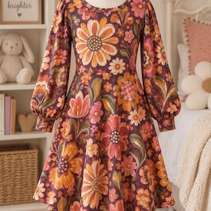 May include: A long-sleeved dress featuring a dark purple base with a vibrant, multi-coloured floral pattern in shades of orange, pink, yellow, and green. The dress has a round neckline, gathered puff sleeves with cuffed wrists, and a flared skirt.