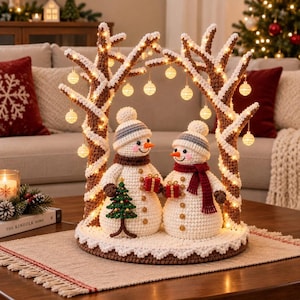 May include: A crochet snowman couple stands under two brown and white yarn trees decorated with glowing string lights and hanging ornaments. The snowmen wear hats and scarves, holding small red gift boxes, with a tiny green tree between them.