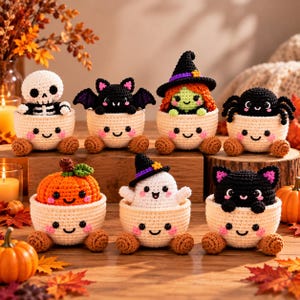 以下が含まれることがあります： 笑顔のクリーム色のカップに入った7体のハロウィーンのかぎ針編み人形。骸骨、黒いコウモリ、緑の髪の魔女、黒いクモ、オレンジ色のカボチャ、白い幽霊、黒い猫のキャラクターが、小さな茶色の足が付いたカップの中にそれぞれ収まっています。