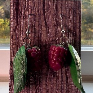 May include: A pair of dangle earrings featuring realistic dark red raspberry charms paired with textured green leaves. The fruit and foliage are suspended from silver-toned metal hooks, creating a detailed botanical design that mimics the appearance of fresh berries.