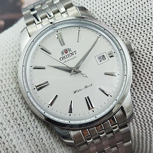 Puede incluir: Un reloj de pulsera automático Orient con caja redonda plateada y esfera blanca con marcadores de hora plateados, manecillas plateadas, una ventana de fecha que muestra el 26 y el texto Orient Automatic y Water Resist, con brazalete metálico.