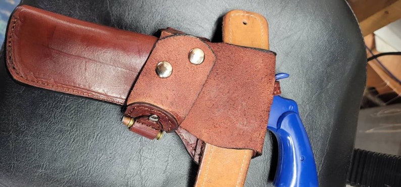 Op de afbeelding: Een bruine leren holster met een blauw plastic speelgoedpistool erin. De holster heeft een klep die het pistool op zijn plaats houdt. De holster is ontworpen om te dragen aan een riem.