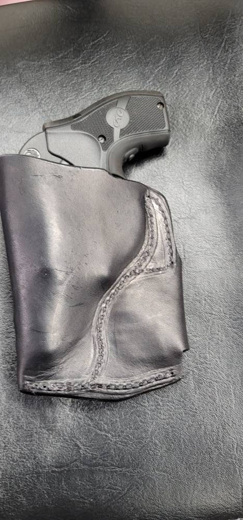 May include: A black leather holster with a stitched design, holding a black handgun with a silver barrel and a black grip. The gun has a silver inscription that reads "380".