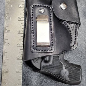 May include: A black leather holster with a metal clip and a black handgun inside. The holster has a flap with a snap closure.