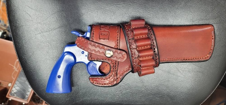 Op de afbeelding: Een bruine leren holster met een blauwe revolver en een lus voor extra munitie. De holster heeft het woord "D.W." erop gegraveerd.