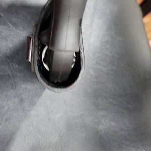 May include: Close-up of a black leather bag with a silver buckle and a black strap. The strap is attached to the bag with a metal loop.