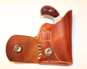Op de afbeelding: Een bruine leren holster met een flap die opengaat om een ​​compartiment voor munitie te onthullen. Een klein zilveren pistool met een houten handvat zit in de holster.