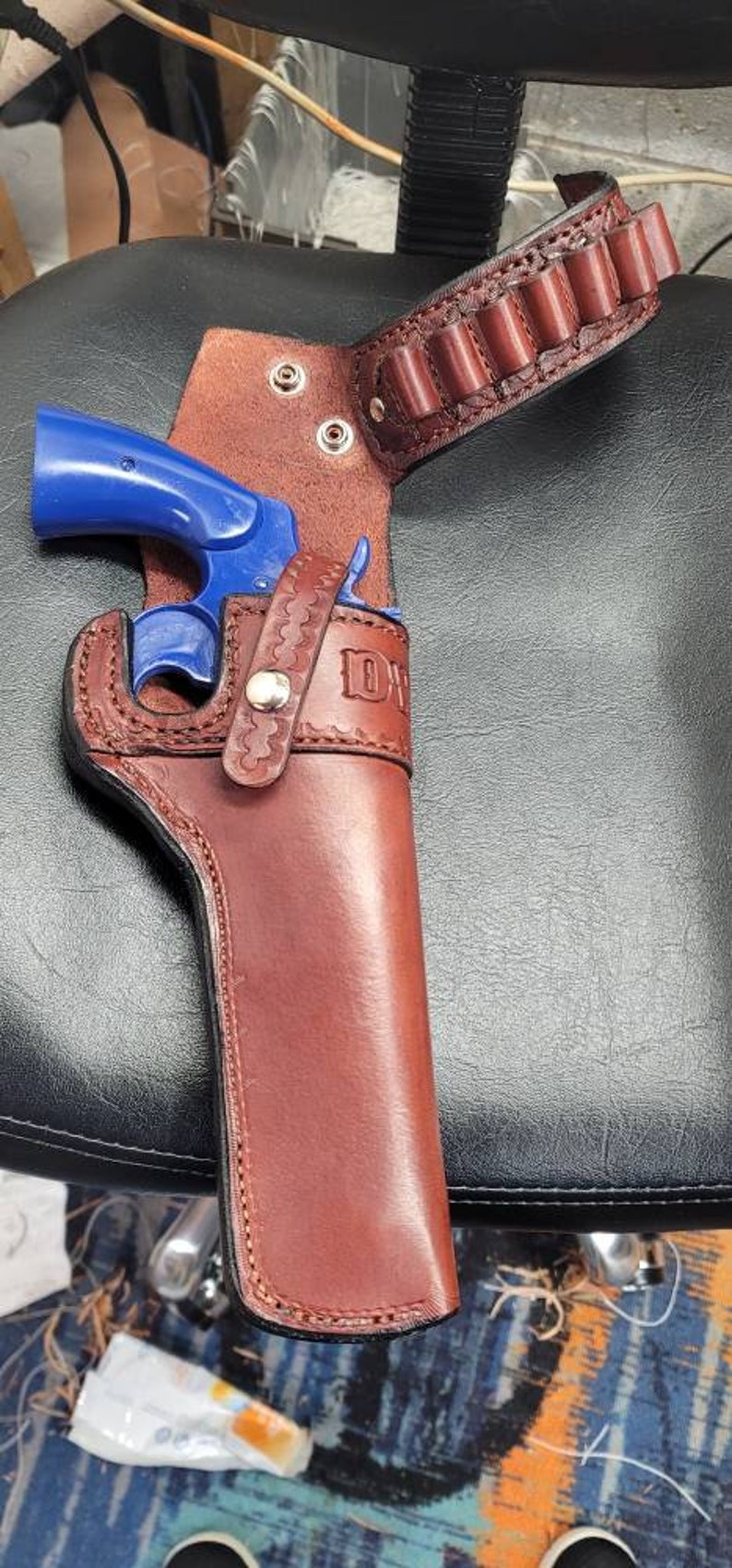 Op de afbeelding: Een bruine leren holster met een blauw speelgoedpistool erin. De holster heeft een riemlus en een decoratief stikselpatroon. De holster is gelabeld "D.W."