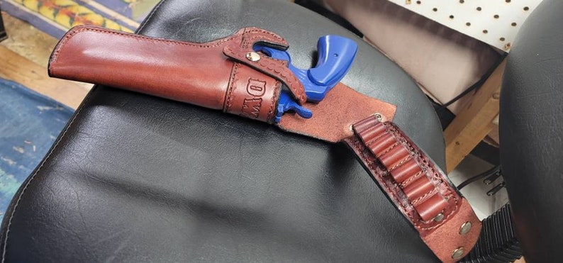 Op de afbeelding: Een bruine leren holster met een blauw speelgoedpistool erin. De holster heeft een riemlus en een magazijnhouder. De holster is gepersonaliseerd met de naam "DAD".