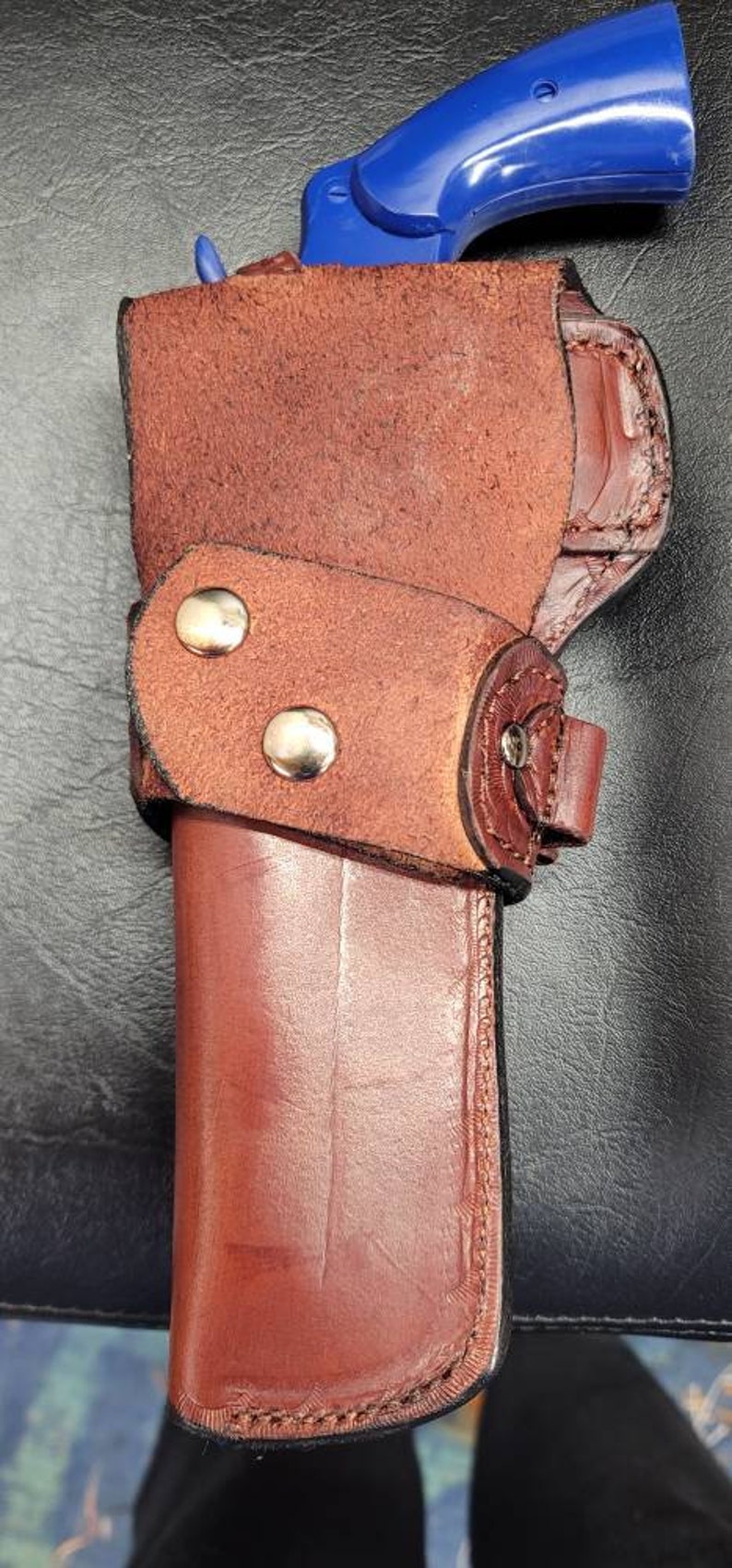 Op de afbeelding: Een bruine leren holster met een blauw plastic pistool erin. De holster heeft een band met twee metalen drukknopen.