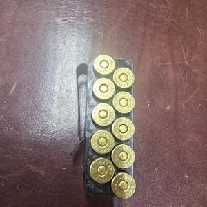 May include: A black 3D-printed ammunition holder featuring a metal belt clip on the side. The rectangular holder contains ten brass-colored cartridges arranged in two vertical rows, with the primer ends facing upward and visible.