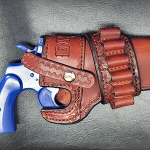 Op de afbeelding: Een bruine leren holster met een blauwe revolver en een lus voor extra munitie. De holster heeft een decoratief stikselpatroon en de tekst "D.W." is in reli&euml;f op de zijkant.