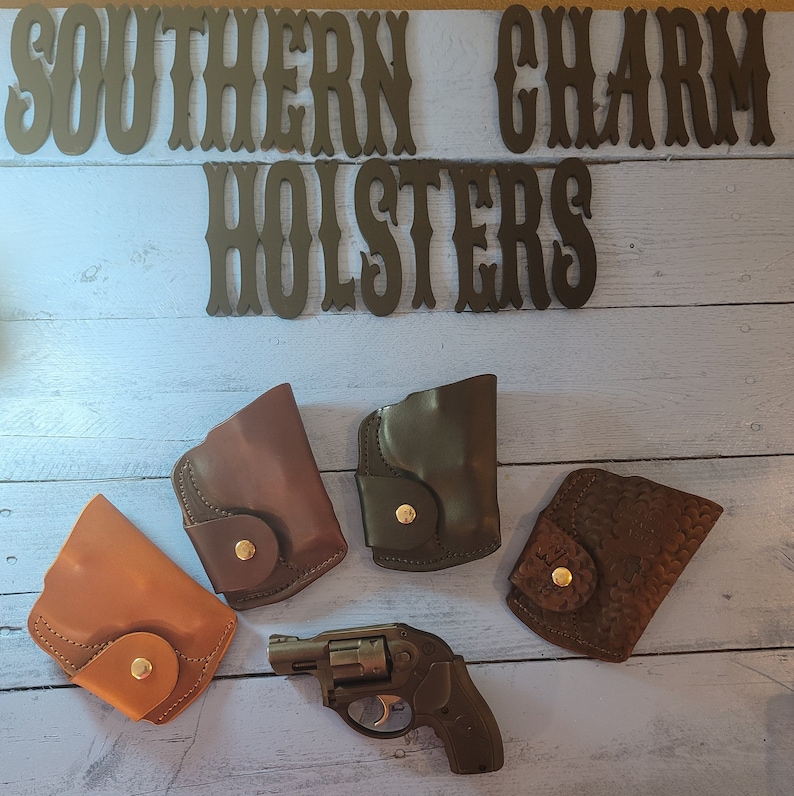 May include: Four different leather holsters in brown, tan, and black, with a small black revolver on a light blue wooden background. The holsters are all closed and have a snap closure. The text "Southern Charm Holsters" is visible at the top of the image.