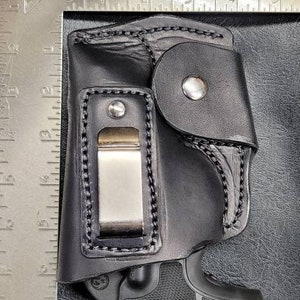 May include: Black leather holster with a clip and a black handgun inside. The holster has a flap with a snap closure.