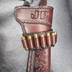 May include: A dark brown leather holster featuring a stamped dragon design and the letters JP. The holster includes an integrated cartridge belt holding six brass-cased bullets and contains a black revolver with a textured grip handle.