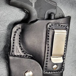 May include: A black leather holster with a metal clip for a handgun. The holster has a stitched design and a smooth finish.
