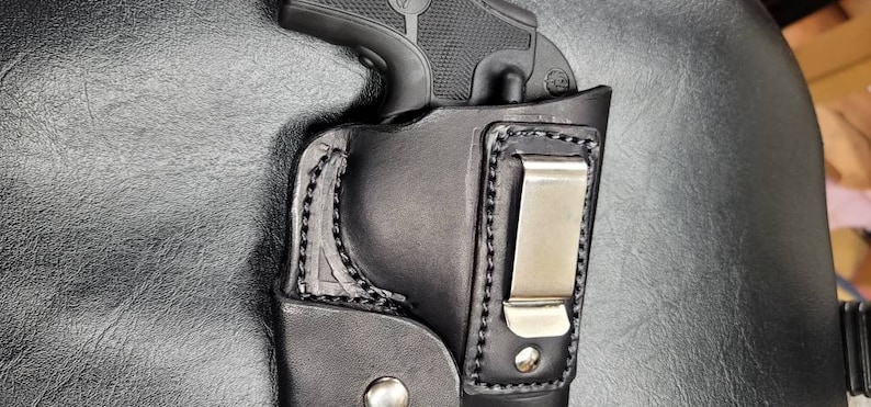 May include: A black leather holster with a metal clip for a handgun. The holster has a stitched design and a smooth finish.