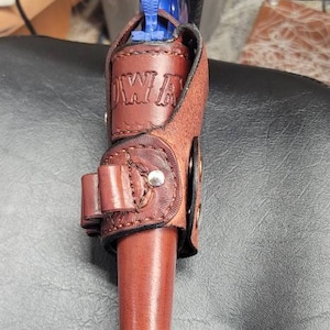 Op de afbeelding: Een bruine leren holster met een blauw speelgoedpistool erin. De holster heeft het woord "COWBOY" erop gegraveerd.