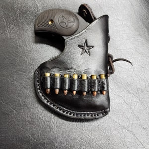 May include: Black leather holster with a star emblem and a row of bullets. The holster holds a black handgun with a star design on the grip. The leather has a textured appearance.