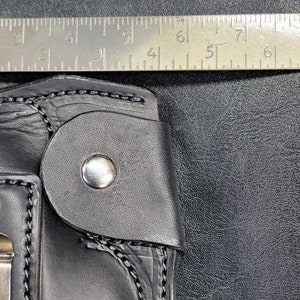 May include: A black leather holster with a silver button and stitching. The holster is designed to hold a handgun. A ruler is visible in the background.
