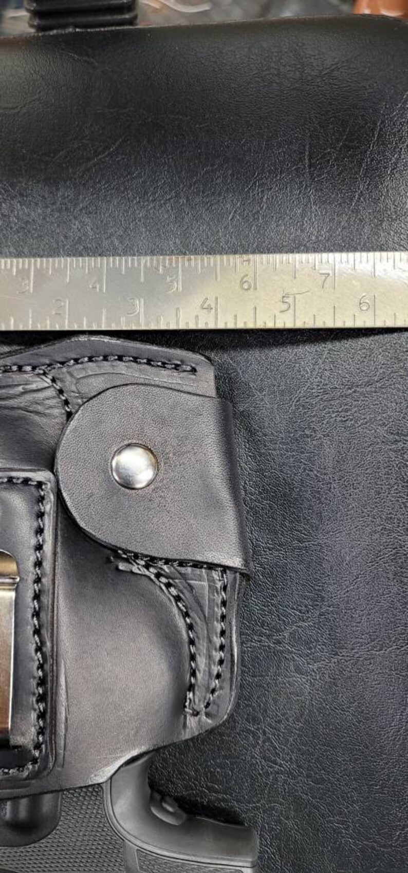 May include: A black leather holster with a silver button and stitching. The holster is designed to hold a handgun. A ruler is visible in the background.