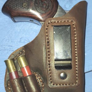 May include: A brown leather holster with a metal clip, holding a revolver with a wooden grip. Two shotgun shells are secured in a leather loop on the side. The holster is designed for concealed carry.