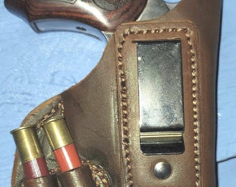 Op de afbeelding: Een bruin leren holster met een metalen clip, met een revolver met een houten handvat. Twee hagelpatronen zijn bevestigd in een leren lus aan de zijkant. Het holster is ontworpen voor verborgen dragen.