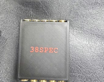 Könnte beinhalten: Ein schwarzes rechteckiges Gerät mit goldfarbenen Anschlüssen. Der Text "38SPEC" ist rot aufgedruckt. Das Gerät hat eine strukturierte Oberfläche und befindet sich vor einem dunklen Hintergrund.