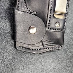 May include: A black leather holster with a silver metal clip and a snap button. The holster is designed to hold a small object, such as a knife or a tool.