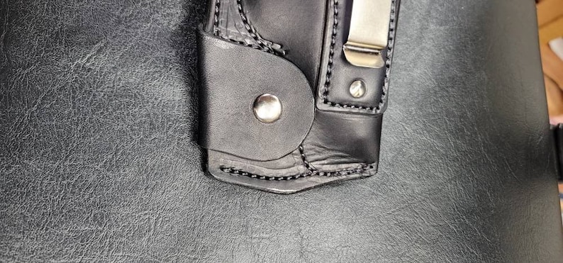 May include: A black leather holster with a silver metal clip and a snap button. The holster is designed to hold a small object, such as a knife or a tool.