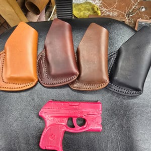 May include: Five leather holsters in different colors, including brown, tan, and black, are displayed on a black surface. A pink plastic toy gun is also shown in the image.