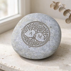 May include: A smooth, light gray river stone featuring a laser-engraved circular design of a tree with intertwined roots and branches. The letters A & M are centered within the tree motif, surrounded by small triangular knotwork patterns.