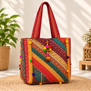 May include: A tote bag featuring a patchwork design of diagonal fabric strips in red, gold, teal, and purple. The bag includes two matching fabric handles, small colorful pom-poms attached to the sides, and a central pom-pom charm.