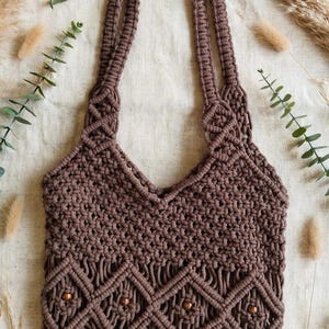 May include: A dark brown macrame tote bag featuring a complex knotted pattern with a central row of diamond shapes accented by small wooden beads. The bag has two integrated shoulder straps and a textured, woven construction throughout the body.
