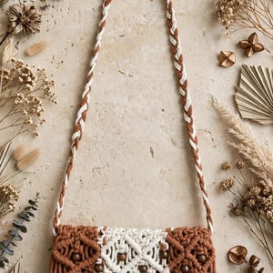 May include: A macrame crossbody bag featuring a braided strap, a rectangular body with a central cream panel flanked by two brown sections, intricate knotted patterns, dark wooden beads, and a long fringe along the bottom edge.