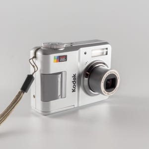 Peut inclure: Un appareil photo numérique Kodak argenté doté d'un objectif rétractable avec l'inscription 36mm-108mm equivalent, un logo Kodak Color Science, un flash intégré et une dragonne grise fixée sur le côté. Le boîtier a une finition métallique.
