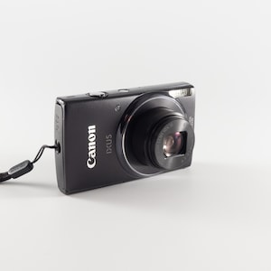 Könnte beinhalten: Eine schwarze Canon IXUS Digitalkamera mit einem markanten Objektivtubus, einem silbernen Auslöser an der Oberseite und einer schwarzen Handschlaufe an der Seite. Auf der Vorderseite sind die Markennamen Canon und IXUS zu sehen.