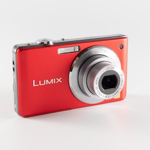 Könnte beinhalten: Eine rote Lumix Digitalkamera mit einem silbernen Leica DC Vario-Elmarit Objektiv mit den Spezifikationen 1:2.8-5.9/5.5-22 ASPH. Das Kameragehäuse verfügt über einen Auslöser an der Oberseite, einen integrierten Blitz und den Markennamen Lumix.