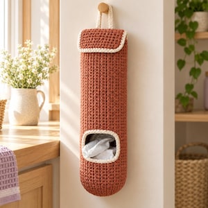May include: A cylindrical, rust-colored crochet bag for storing plastic bags, featuring a top flap, a hanging loop, and a rectangular opening at the bottom with a cream-colored trim, suspended from a wooden wall hook.