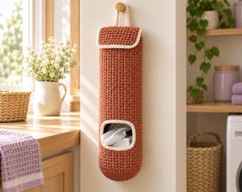 May include: A cylindrical, rust-colored crochet bag for storing plastic bags, featuring a top flap, a hanging loop, and a rectangular opening at the bottom with a cream-colored trim, suspended from a wooden wall hook.