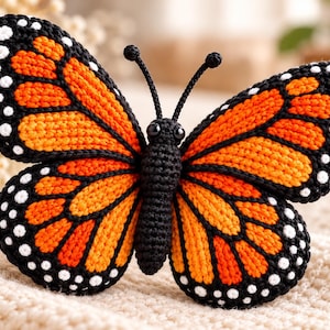 May include: A crocheted monarch butterfly with vibrant orange and black wings featuring white spots along the edges. The butterfly has a textured black body, two thin antennae with rounded tips, and small black bead eyes, crafted with detailed yarn stitches.