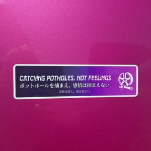 Può includere: Un adesivo rettangolare con sfondo sfumato dal viola al nero su una superficie magenta. Presenta il testo bianco "CATCHING POTHOLES, NOT FEELINGS" sopra caratteri giapponesi e una grafica bianca di una ruota d'auto e una chiave inglese.