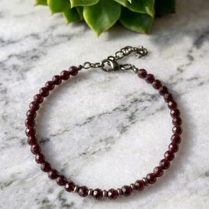 May include: A delicate bracelet featuring small, faceted, deep red garnet beads strung together with dark metallic spacers. The piece includes a dark metal lobster claw clasp and an adjustable chain link extension for a secure fit.