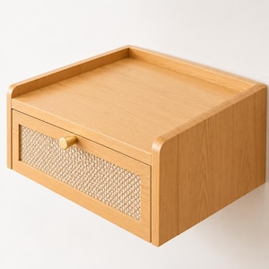 May include: A wall-mounted floating bedside table crafted from light-toned wood, featuring a single drawer with a woven cane front panel and a small, round, gold-coloured metal knob. The top surface has a raised edge on three sides.