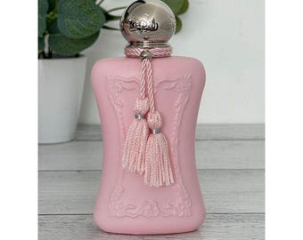 May include: A matte light pink perfume bottle with a curved silhouette and a silver metallic cap. The neck of the bottle is adorned with a pink braided cord featuring two matching tassels. The front surface displays a raised, ornate frame.