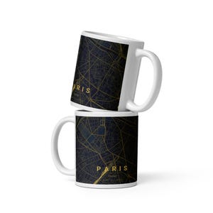 May include: Two white ceramic mugs stacked vertically, each featuring a dark navy blue map of Paris, France, with gold street lines. The text PARIS FRANCE is printed in gold lettering on the front of each mug, along with coordinates.