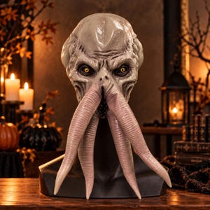 Op de afbeelding: Een volledig hoofdmasker met een bleek, getextureerd wezenontwerp met diepliggende gele ogen, een prominent voorhoofd en vier lange, naar beneden hangende tentakels die de mond bedekken, getoond op een zwarte etalagepopkop.
