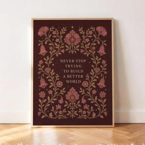 May include: A rectangular wall art print in a light wood frame featuring a dark maroon background with gold floral vines and the text NEVER STOP TRYING TO BUILD A BETTER WORLD centered in a serif font surrounded by small star accents.