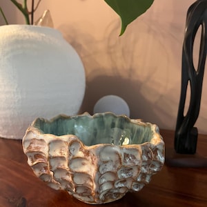 Puede incluir: Un pequeño cuenco de cerámica redondo con un exterior texturizado tipo panal, con un esmalte moteado en tonos marrón y crema. El interior del cuenco tiene un acabado de esmalte verde claro liso y brillante, que contrasta con la superficie exterior.