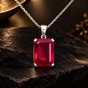 May include: A rectangular, deep red gemstone pendant set in a silver-toned metal prong setting, suspended from a delicate silver-toned chain. The pendant features a faceted cut that reflects light, showcasing a vibrant, saturated crimson hue.